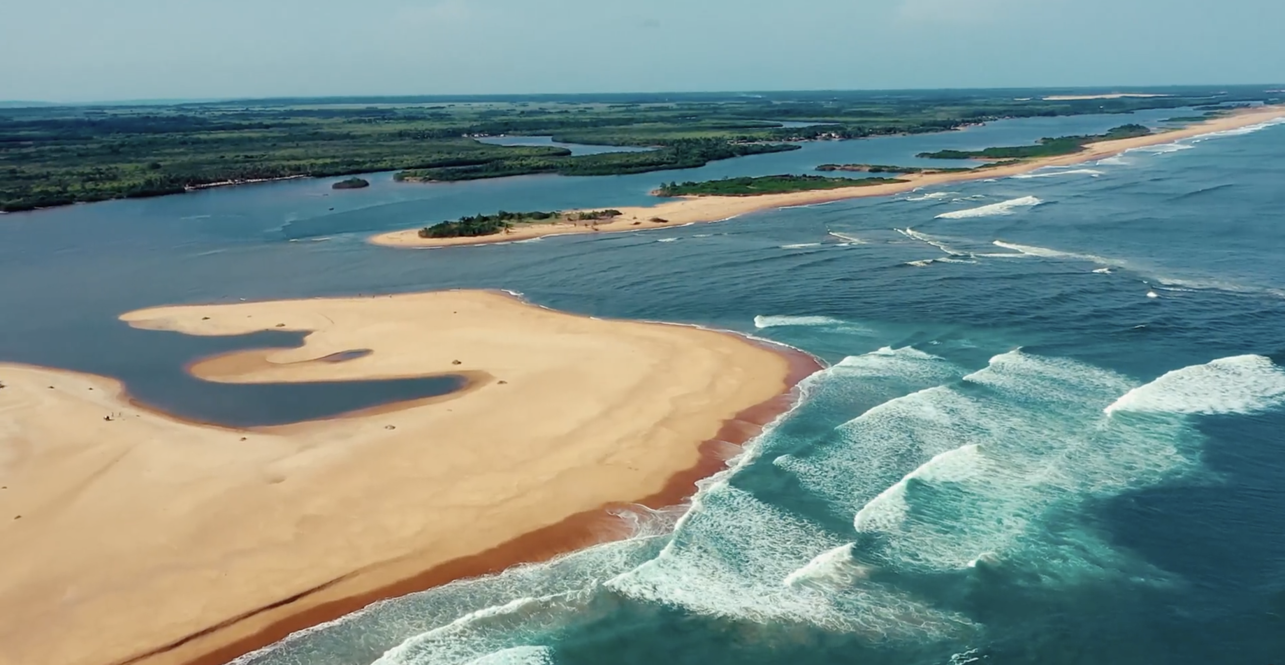 Vue aérienne de la cité balnéaire du Bénin de sable où les vagues viennent se briser sur le rivage, avec des îles verdoyantes en arrière-plan sous un ciel dégagé.