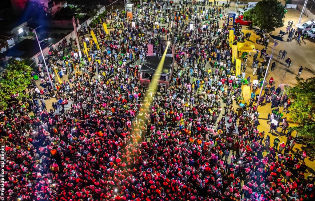 Une foule nombreuse se rassemble en plein air, la nuit, autour d'une scène éclairée par des lumières vives et des bannières colorées, vue d'en haut.