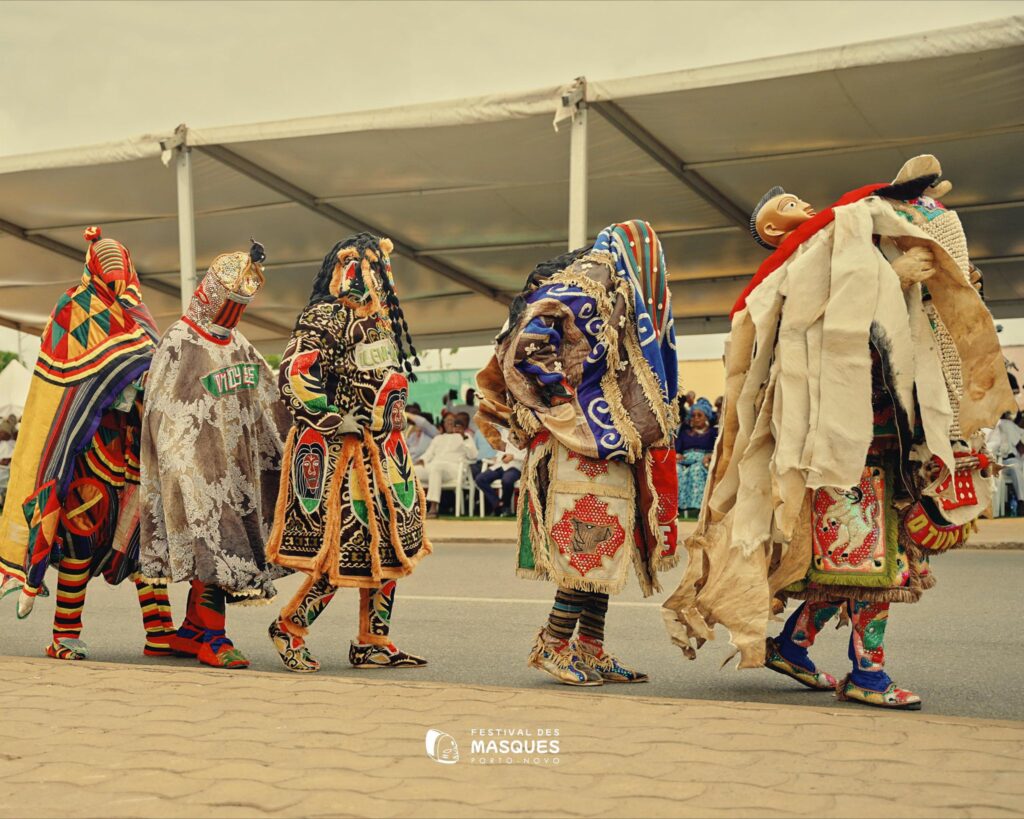Cinq personnes portant des masques et des costumes traditionnels élaborés et colorés marchent en ligne lors d'un festival culturel, avec des tentes et des spectateurs assis à l'arrière-plan.