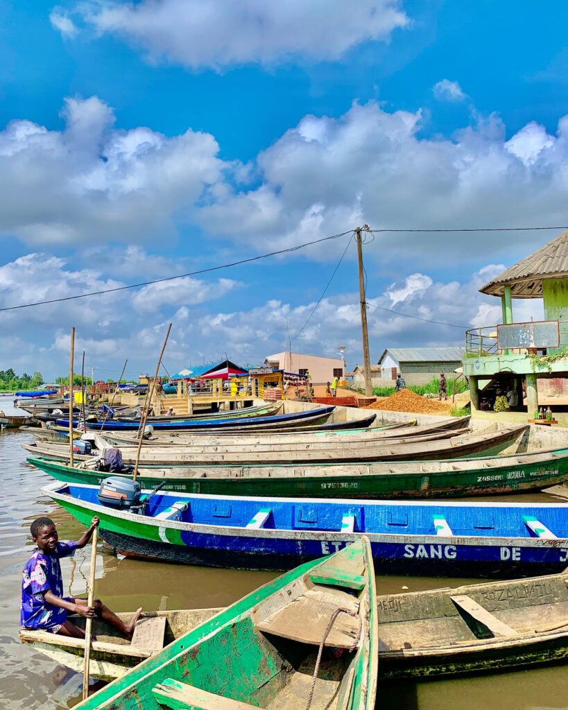 Jeune garçon pagayant une pirogue près de l'embarcadère menant à Ganvié au Bénin , entouré de barques colorées et de maisons en bord de lagune sous un ciel bleu nuageux.