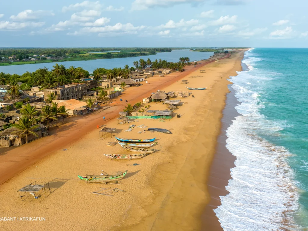 Vue aérienne de la plage d'Avlo à Grand-Popo au Bénin, de sable avec des bateaux de pêche sur le rivage, des vagues de l'océan et un village voisin avec des palmiers et des bâtiments simples.