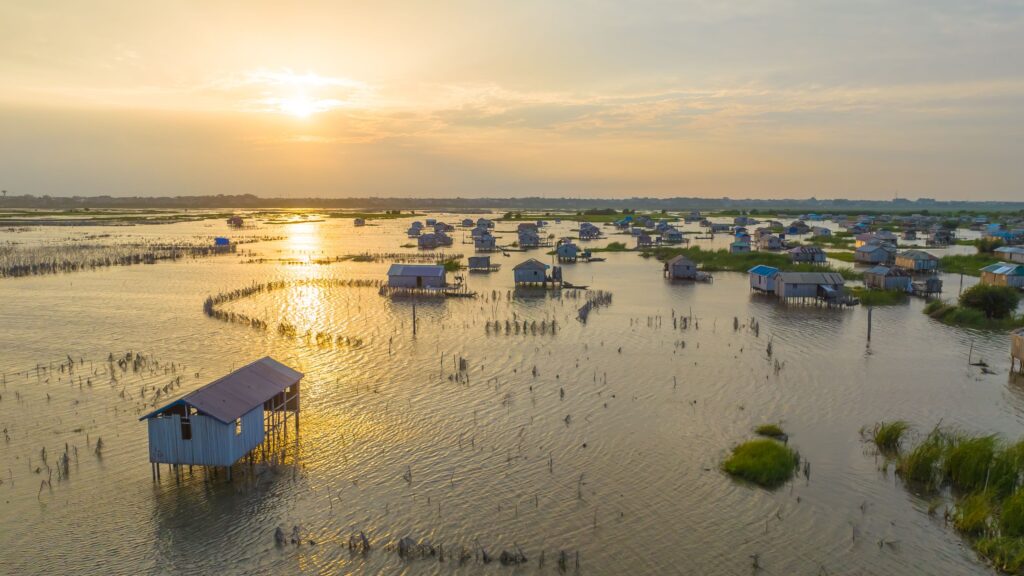 Vue aérienne de maisons sur pilotis dans la cité lacustre de Ganvié au Bénin au coucher du soleil, avec une végétation éparse et une eau calme.