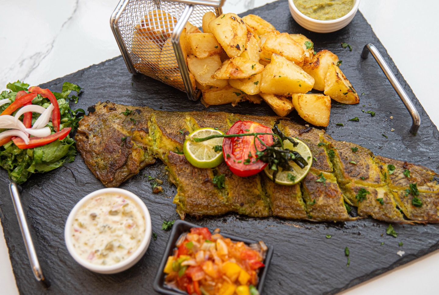 Poisson grillé garni de citron vert et de tomates, servi avec des pommes de terre rôties, une salade, une sauce verte, un dip crémeux et une salsa de légumes en dés sur une assiette en ardoise noire.