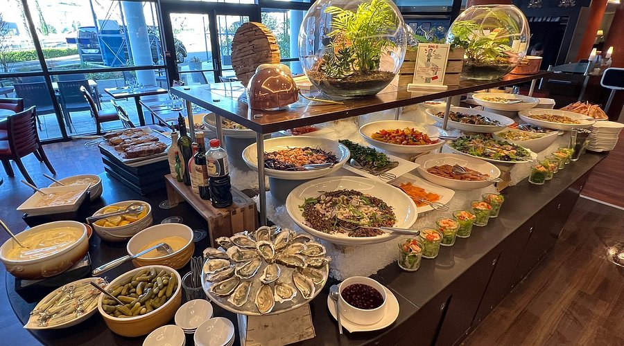 Une table de buffet présente un assortiment de salades, d'huîtres, de sauces, de pain, de cornichons et de petits bocaux d'amuse-gueule, surmontés de terrariums décoratifs en verre.