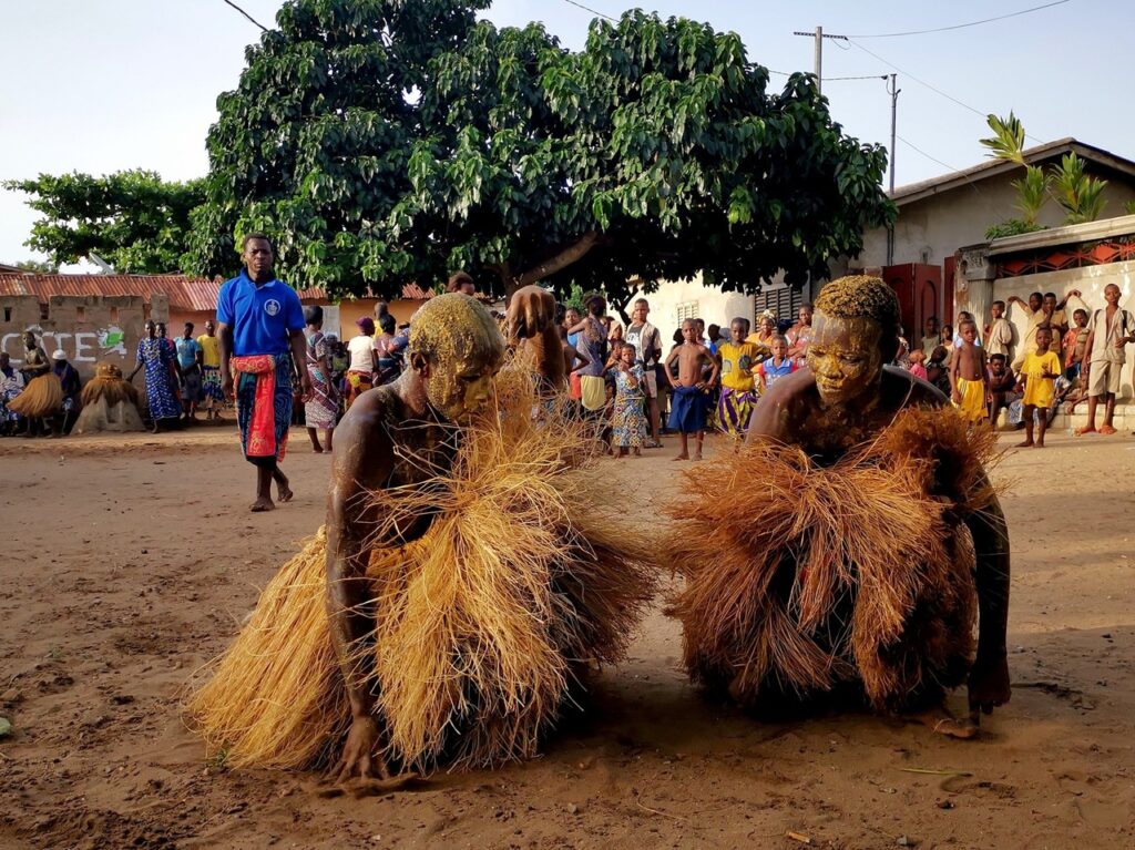 Deux adeptes du vodoun au Bénin vêtues de costumes de paille sont accroupies sur un sol sablonneux, entourées d'une foule de spectateurs dans un village en plein air avec des arbres et des bâtiments en arrière-plan.
