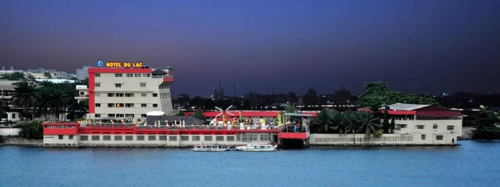 Un bâtiment de plusieurs étages portant le nom d'"Hôtel du Lac", avec des accents rouges et blancs, se trouve au bord de l'eau au crépuscule, avec des bateaux amarrés à proximité et le paysage urbain à l'arrière-plan.