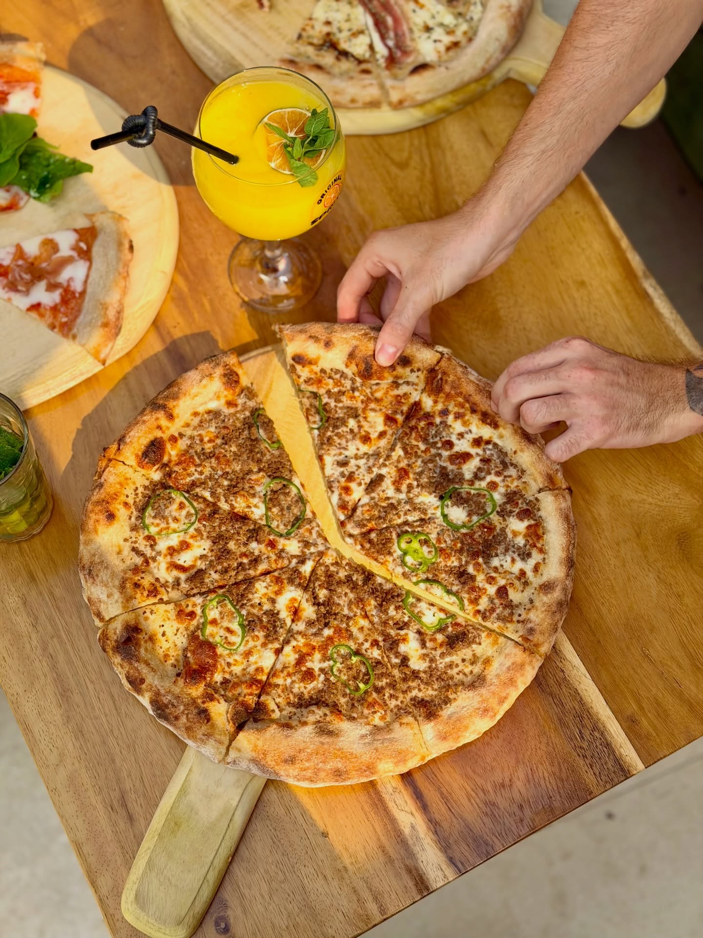 Une personne saisit une part de pizza garnie de viande hachée et de piments jalapeños sur une planche en bois, avec une boisson jaune et d'autres pizzas à proximité sur la table.