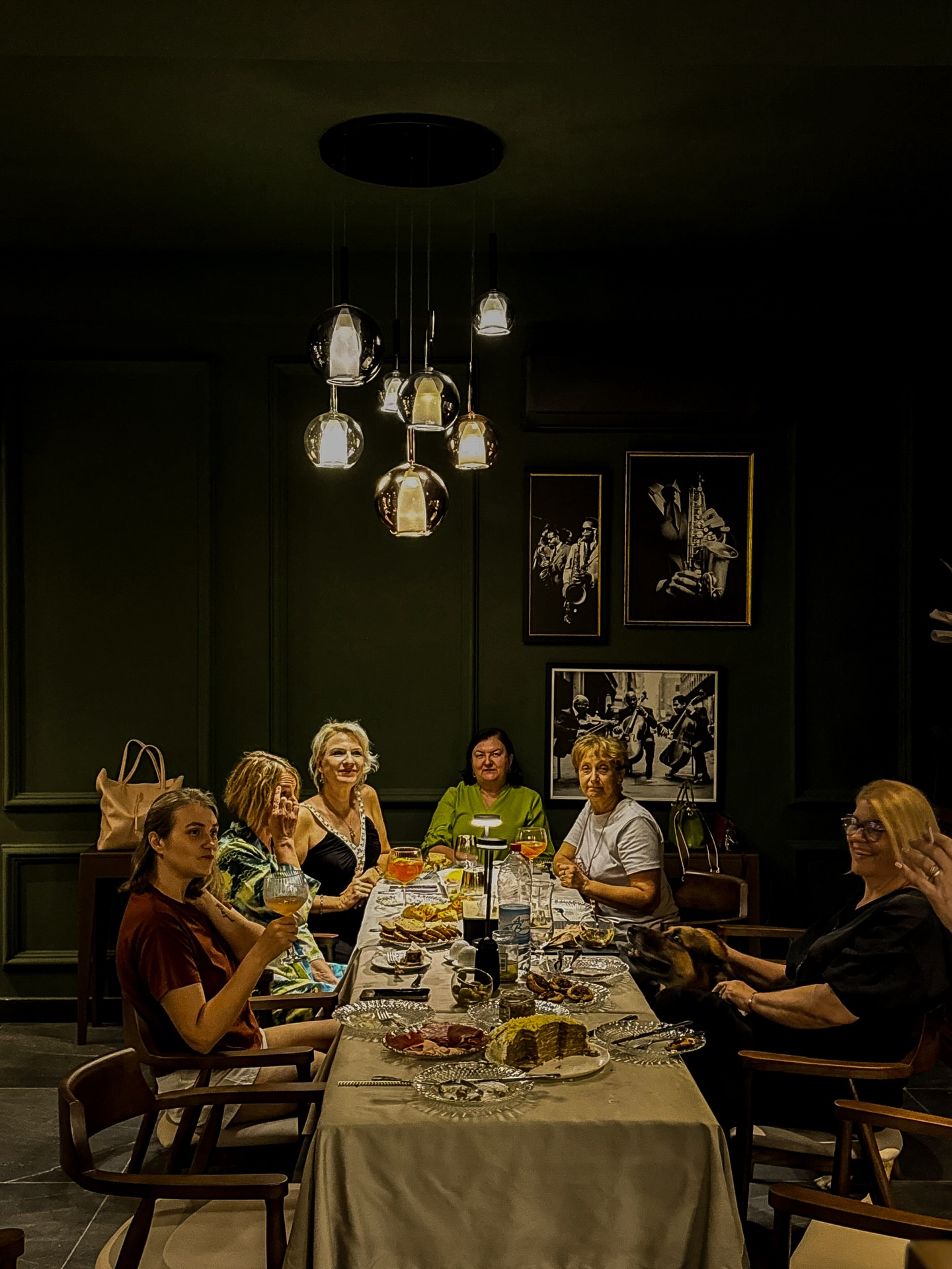Six femmes sont assises autour d'une longue table avec de la nourriture et des boissons, dans une pièce faiblement éclairée aux murs sombres et aux photos encadrées.