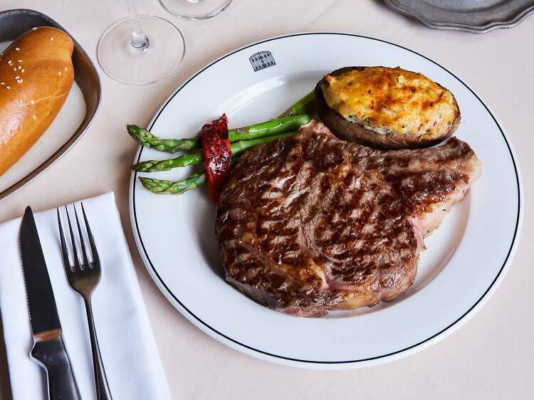 Un steak grillé servi avec une pomme de terre cuite deux fois, des asperges et un poivron rouge rôti sur une assiette blanche, avec un petit pain, des ustensiles et un verre de vin à proximité.