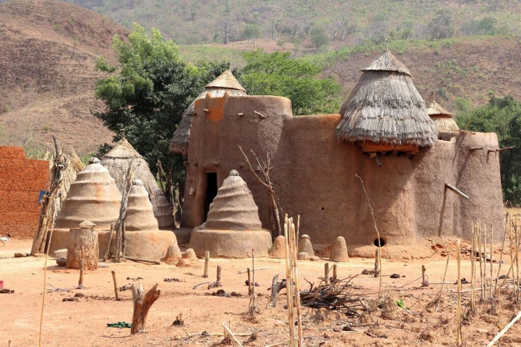 Structures traditionnelles en terre et en chaume avec des toits coniques dans un paysage rural avec un sol sec et des collines en arrière-plan.